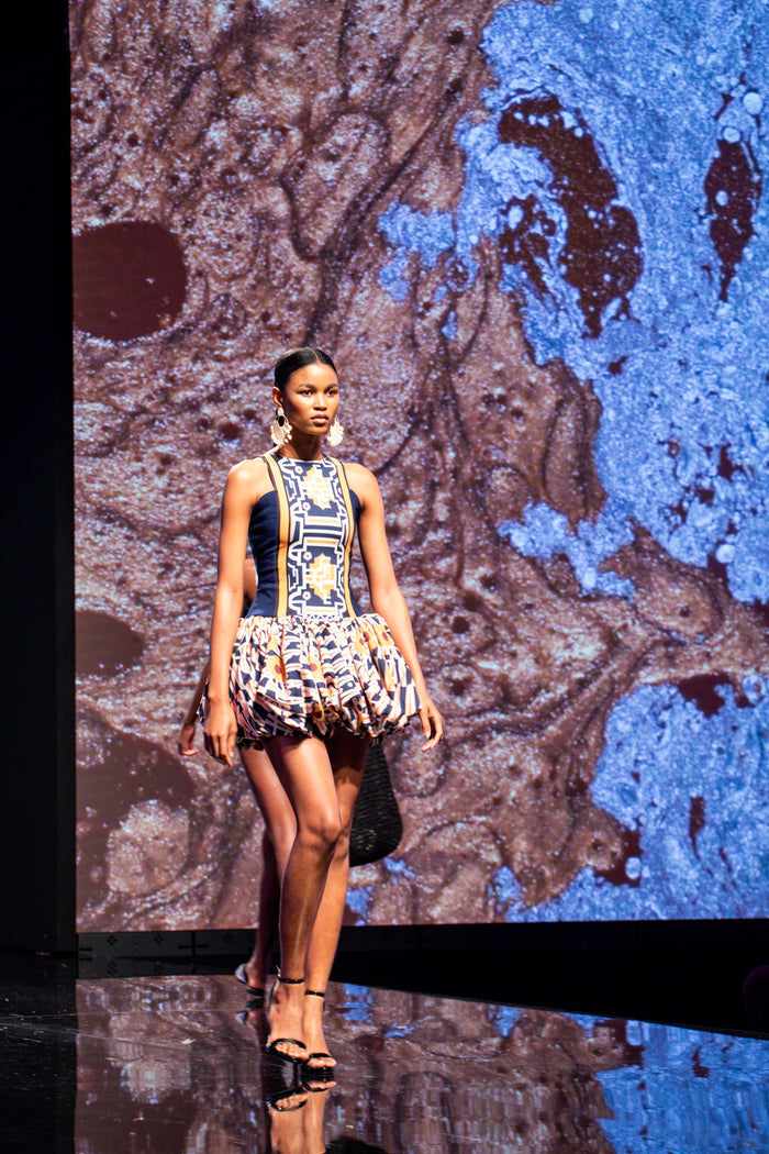 A stylish model wearing a vibrant Fabrosanz bubble hem mini dress with bold blue, mustard yellow, and geometric tribal-inspired prints, walking down the runway at a 13th annual Durban Fashion Fair.