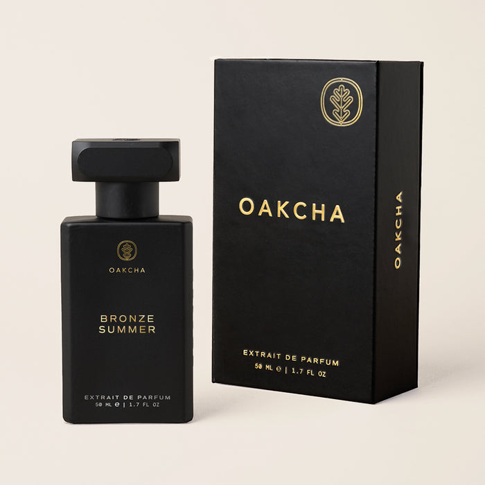 Front view of Bronze Summer 50ml perfume bottle and black box with gold logo on beige backdrop