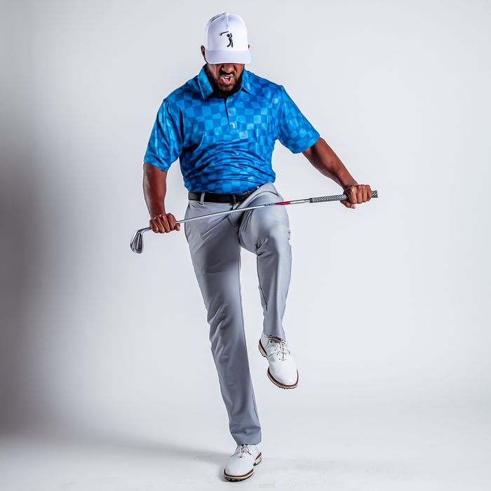 Wearing the Checked Out - Polo, an athletic-fit blue checkered golf shirt with UPF 50 protection, plus light gray pants and white shoes, a man steps forward gripping a golf club, his focused expression set against a plain white background.