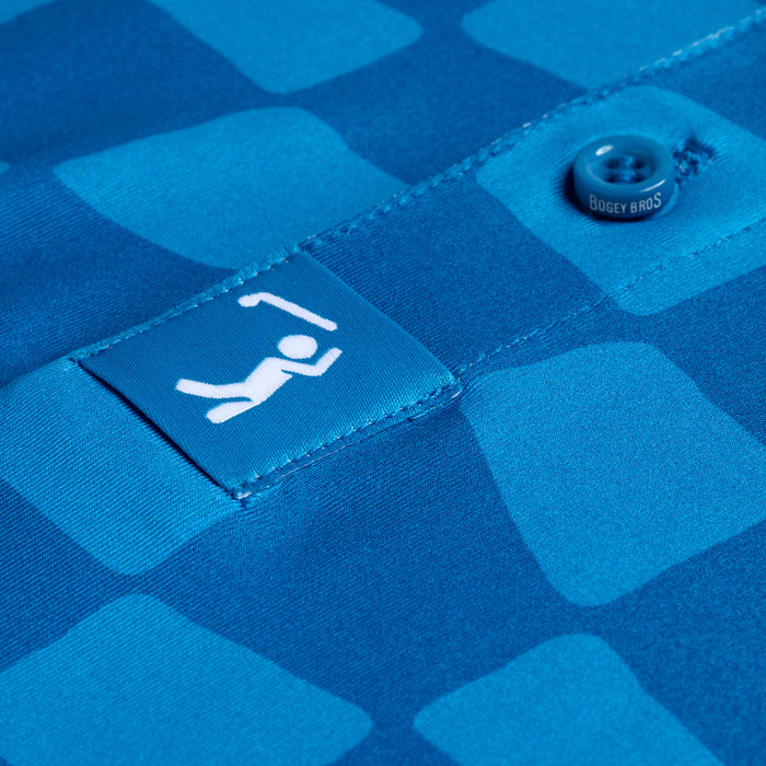 Close-up of the Checked Out - Polo, showcasing blue fabric with light blue mosaic squares. Features a tag with a stick figure under a golf flag, "BOGEY BROS" button, athletic fit, quality stitching, and built-in UPF 50 protection.