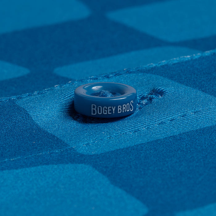 Close-up of a glossy "BOGEY BROS." button on the Checked Out - Polo, sewn onto blue fabric with geometric patterns. The athletic fit and visible stitching showcase the polo’s quality construction.