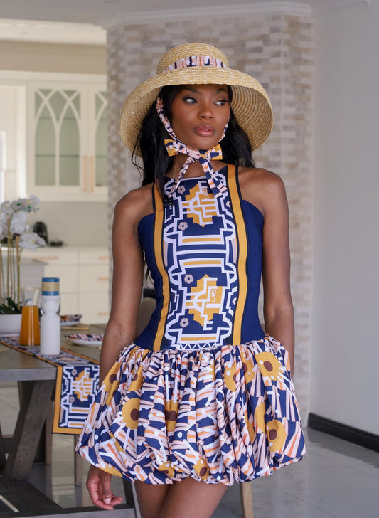 A stylish woman wearing a vibrant Fabrosanz bubble hem mini dress with bold blue, mustard yellow, and geometric tribal-inspired prints, paired with a chic straw hat featuring matching Tshwane print fabric ties, standing in a bright modern room.