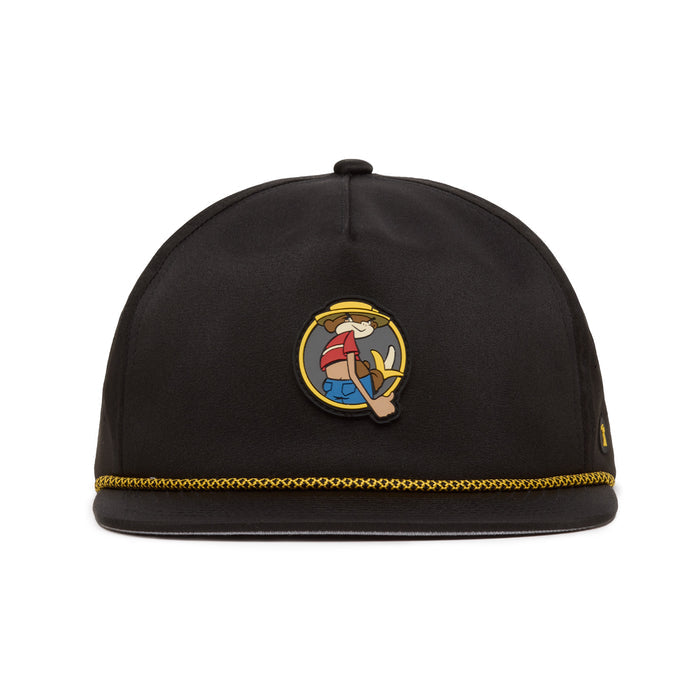 A black hat with gold trim at the brim features a cartoon character patch on the front, showing a smiling person in a hat holding up three fingers. The hats material appears smooth, and its shape is rounded without a visible brim.