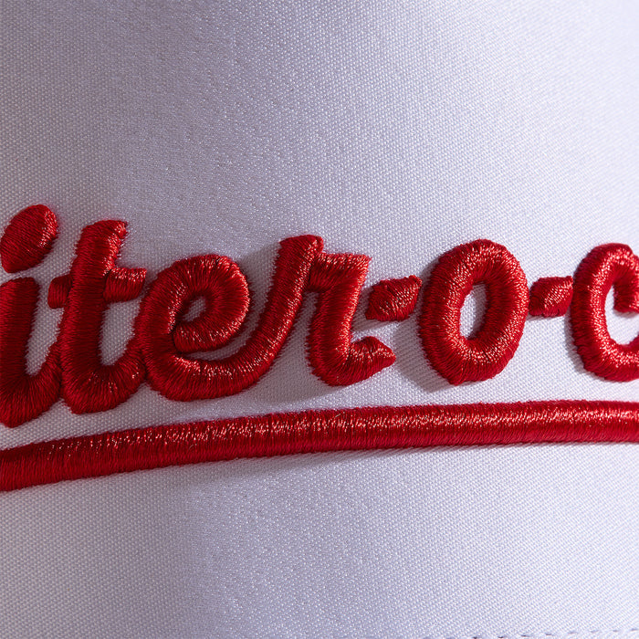 Close-up of red script embroidery on white fabric, showing the text iter-o- with bold, raised stitching. A red curved underline runs beneath the letters. The texture of the thread and fabric weave is clearly visible.