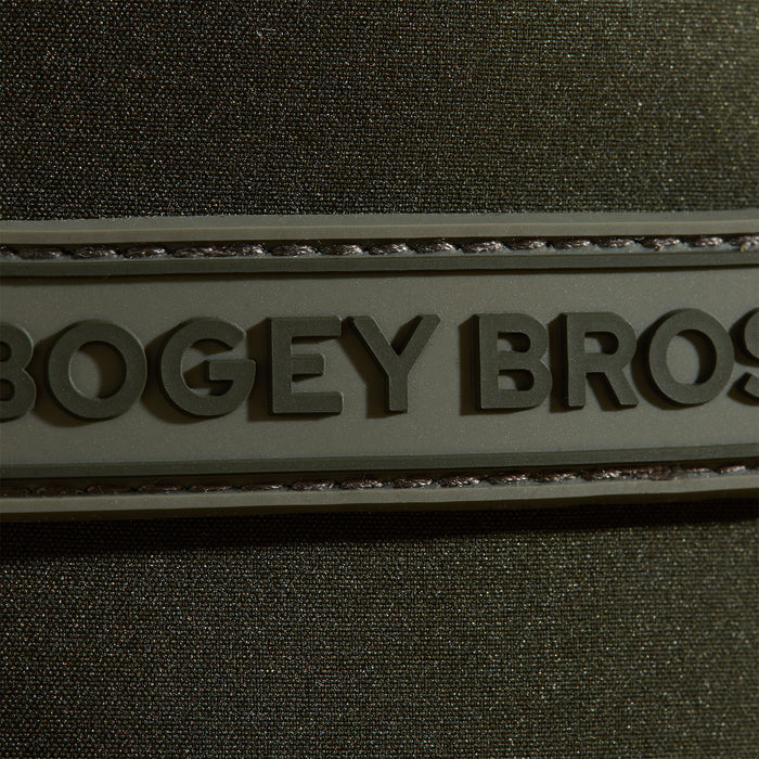 A close-up of a textured dark green fabric with a raised rubber patch that has the embossed, partially visible text BOGEY BROS. The patch is stitched onto the fabric, and the lighting highlights the materials texture and details.