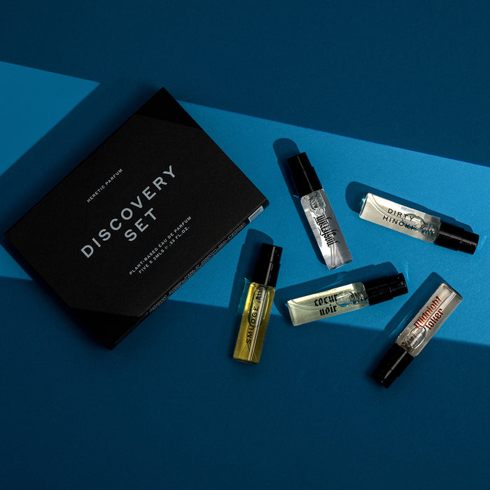 A perfume discovery set featuring a black box labeled 'DISCOVERY SET' and five small glass spray bottles with black caps, arranged on a blue background with dramatic shadows.
