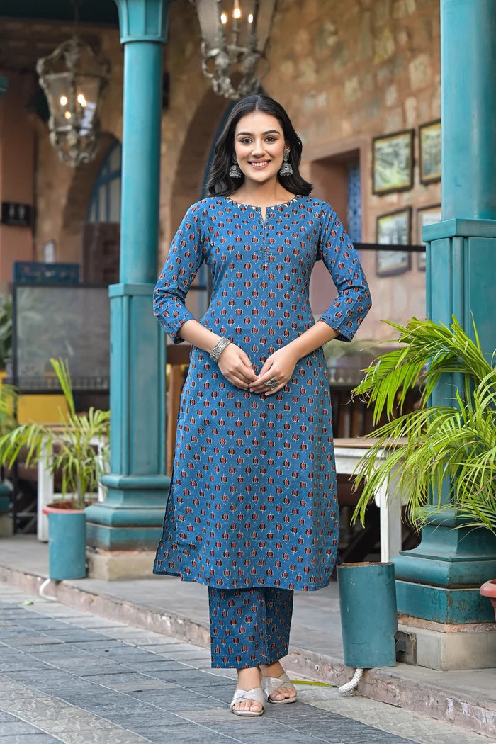 Woman in a blue patterned outfit standing outdoors with plants and architectural elements in the background