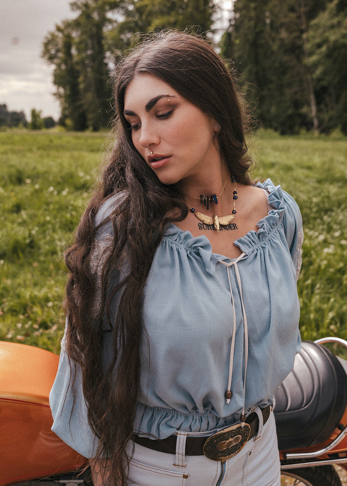 Woman in a light blue blouse by Soul Sister since 1969 sitting on a motorcycle in a grassy field with trees in the background