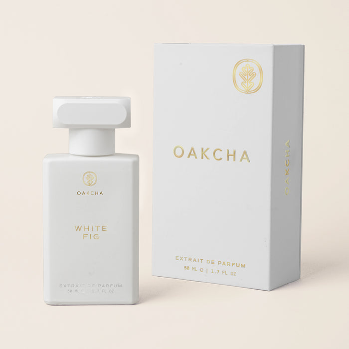 Front view of White Fig perfume bottle with frosted white bottle and gold OAKCHA logo beside box on beige background