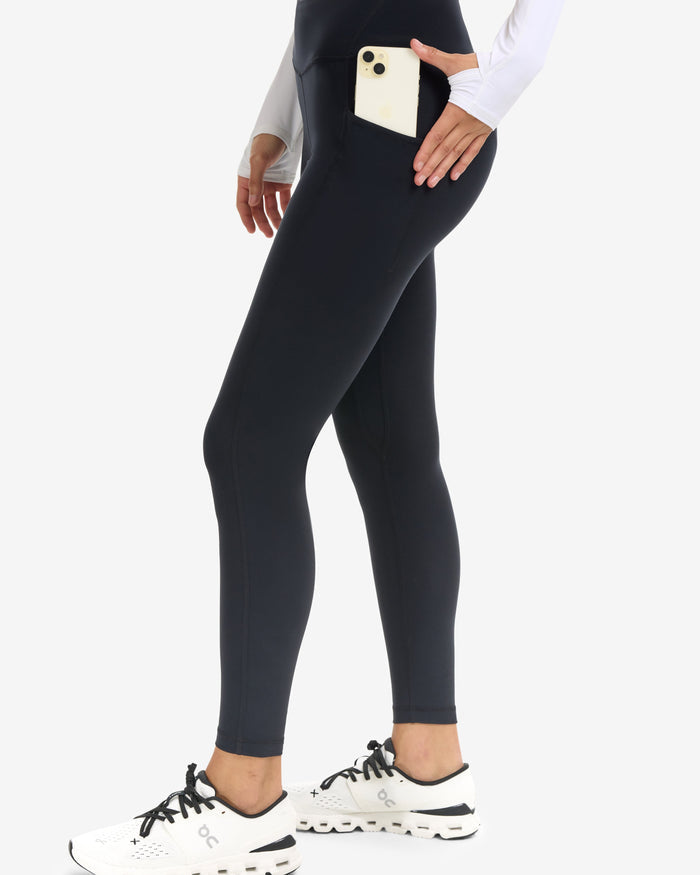 Person wearing black leggings with a pocket, white sneakers, and a white long-sleeve shirt on a white background.